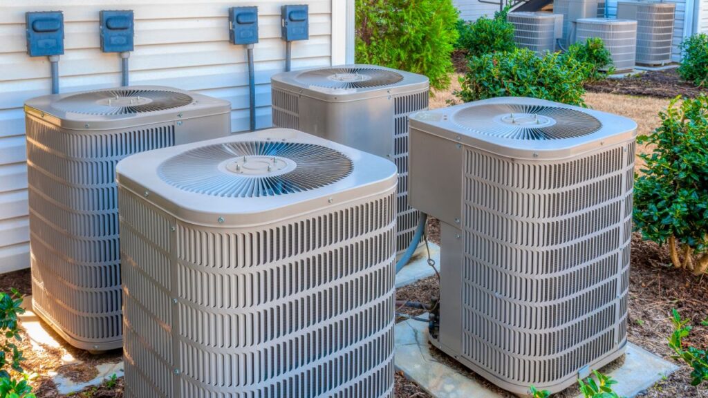 Four large outdoor air conditioning units sit on concrete pads beside a building, surrounded by electrical boxes, shrubs, and a grassy area.
