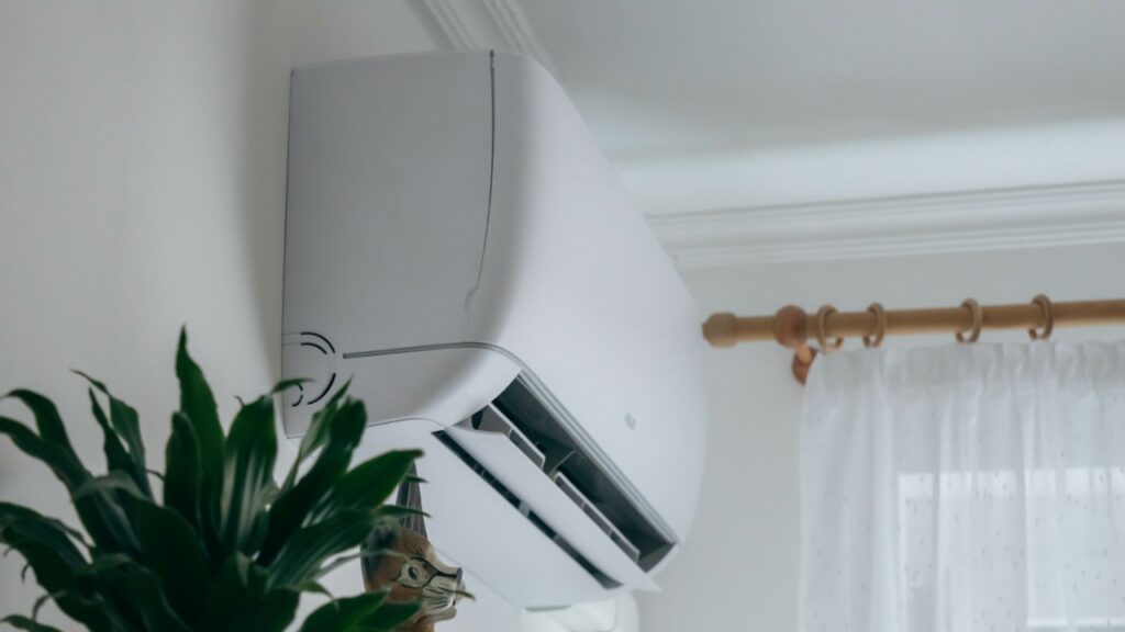 A white wall-mounted air conditioner is installed above a plant near a window with sheer curtains and a brown curtain rod.
