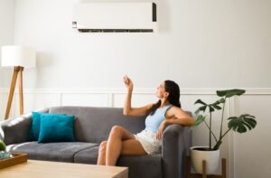 Woman sitting on a gray couch uses a remote control to operate a wall-mounted air conditioner in a modern living room.